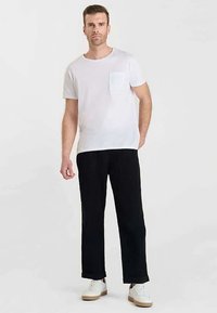 Camiseta blanca de algodón con bolsillo en el pecho, combinada con pantalones negros de algodón. El modelo está de pie con zapatillas blancas sobre un fondo neutro.