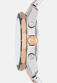 Hõbedane ja roosakuldne kell ümmarguse korpuse, kaheksanurkse raamiga, tekstuuriga nupudega ja ümmarguse krooniga, millel on tume valge.