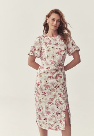 Kobieta o falowanych włosach, ubrana w białą sukienkę midi z różowym kwiatowym nadrukiem, bufkami i rozcięciem z przodu, stojąca z rękami z tyłu.