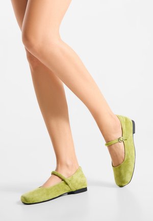 Grüne Wildlederflats mit Riemen- und Schnallendetail, rundem Zeh, niedrigem Absatz und glatter Textur, präsentiert an einem Bein.