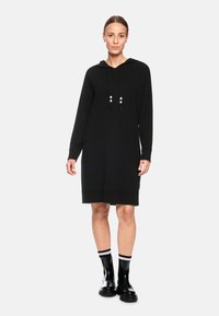 Schwarzes gestricktes Kleid mit langen Ärmeln und Kapuze, ausgestattet mit einem gerippten Saum und verstellbaren Kordeln. Kombiniert mit glänzenden schwarzen Ankle Boots.