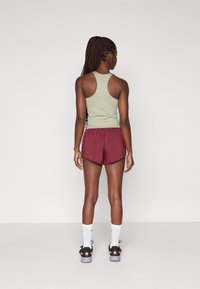 Idrottare iklädd en ljusgrön ärmlös topp och marsalaburna shorts, med svarta och lila sneakers och vita strumpor. Strukturert hår synligt.