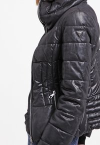 Schwarze gepolsterte Jacke mit glänzender, lederähnlicher Textur, hohem Kragen, diagonalem Reißverschluss und dreieckigem Logo-Akzent auf der Schulter.