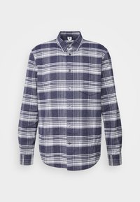 Chemise à manches longues boutonnée avec motif à carreaux bleu marine et blanc, col et poche poitrine sur fond clair uni.