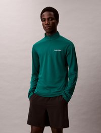 Dlhý rukáv smaragdovo zelený top s kvartovým zipsom, s malým bielym logom Calvin Klein. V kombinácii s čiernymi šortkami, hladká textúra.