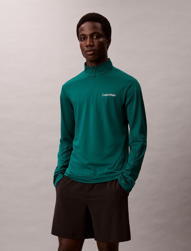 Dlhý rukáv smaragdovo zelený top s kvartovým zipsom, s malým bielym logom Calvin Klein. V kombinácii s čiernymi šortkami, hladká textúra.