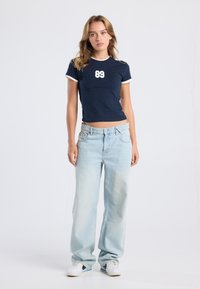 Navy short-sleeve top with white accents and "89" graphic; paired with light blue wide-leg jeans and white sneakers.
