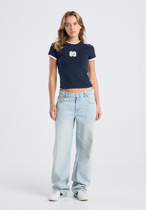 Marineblauw korthangend shirt met witte accenten en een "89" graphic; gecombineerd met lichtblauwe wijde jeans en witte sneakers.
