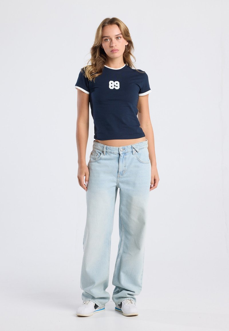 Navy short-sleeve top with white accents and "89" graphic; paired with light blue wide-leg jeans and white sneakers.