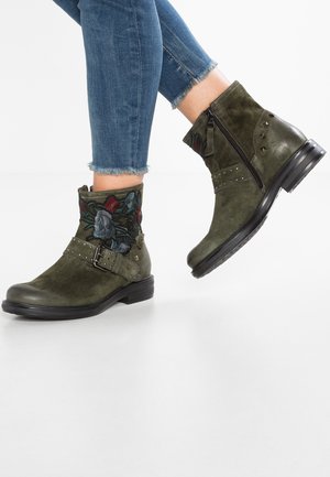 Grüne Wildleder-Stiefeletten mit Blumenstickerei, seitlichem Reißverschluss, Schnallenriemen und Nieten-Details, kombiniert mit blauem Jeans mit ausgefranstem Saum.