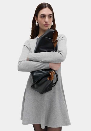 Sort lædertaske med et blødt, krøllet design, med en brun inderside og tynde stropper. Modellen har en lysegrå kjole på.