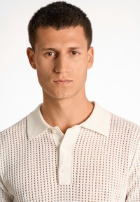 Stickad polo i krämfärg med öppen väv, med krage och knappslå med tre knappar, slät textur och fina horisontella ränder.