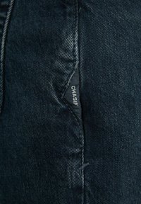 Gros plan sur un tissu denim bleu foncé avec une couture visible et une petite étiquette noire portant l'inscription « "CHASIN" » cousue dans la couture.