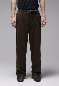 Pantalon marron, ample et à la texture lisse, avec une ceinture noire et un détail de chaîne. Porté avec des chaussures noires et un haut noir.