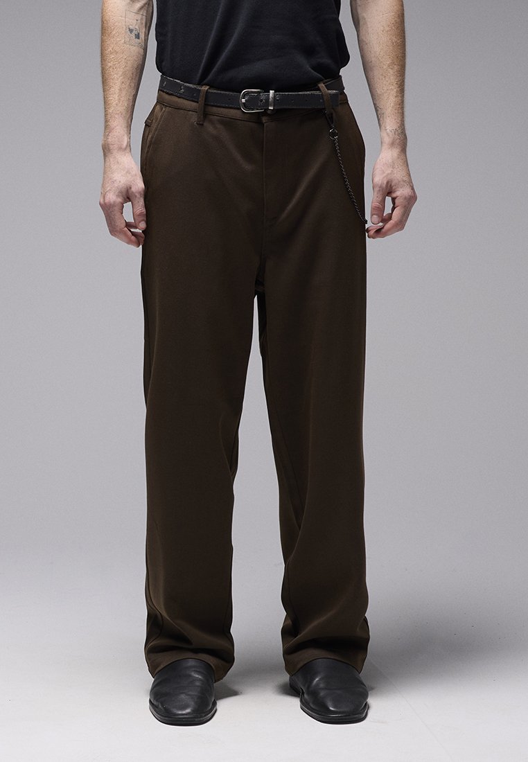 Pantalon marron, ample et à la texture lisse, avec une ceinture noire et un détail de chaîne. Porté avec des chaussures noires et un haut noir.