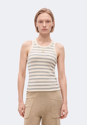 Une femme se tient devant un fond uni, vêtue d’un haut sans manches à rayures, d’un pantalon beige à jambes larges avec des poches et de chaussures noires.