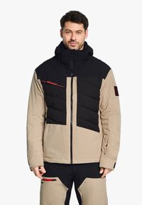 Doudoune avec un schéma de couleurs beige et noir, présentant une texture matelassée sur la poitrine, la capuche et la fermeture éclair ; accents de logo en rouge.