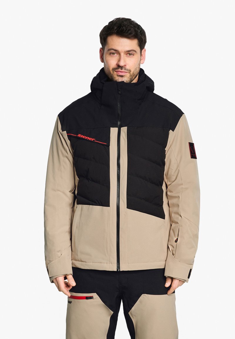 Doudoune avec un schéma de couleurs beige et noir, présentant une texture matelassée sur la poitrine, la capuche et la fermeture éclair ; accents de logo en rouge.