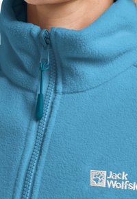 Giacca in pile azzurro chiaro con colletto alto, zip frontale e logo visibile sul petto. Texture morbida e finitura liscia.