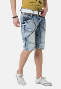 Jeansshorts in einem verblassten Blau mit diagonalen Nähmustern. Abgerundet mit einem weißen Gürtel und getragen mit weißen Turnschuhen.
