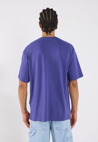 Violets koka t-krekls ar brīvu piegriezumu, īsām piedurknēm un apkakli ar apaļu izgriezumu. Tam ir vienkārša dizaina, bez redzamām logo vai rakstiem.