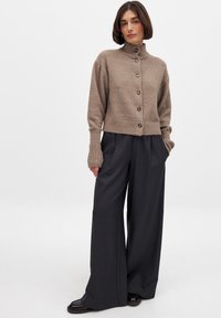 Beige neulecardigan, jossa on korkea kaulus, napitti etuosa ja ribbauskut, yhdistettynä löysiin tummanharmaisiin housuihin ja tasapohjaisiin kenkiin.