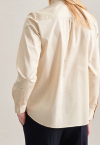 Light beige button-up shirt with long sleeves, back pleat detail, rounded hem, and cuff buttons. Smooth fabric texture, tailored fit.