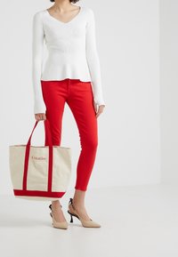 Top blanco de manga larga con canalé, jeans ajustados rojos y zapatos de tacón beige. Bolsa de lona con asas rojas y la palabra "Creativo" bordada en rojo.