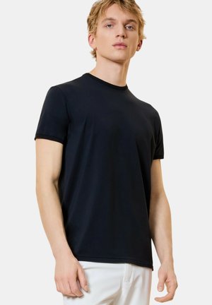 Giovane uomo con capelli biondi che indossa una maglietta nera aderente a maniche corte e pantaloni bianchi, in piedi contro uno sfondo bianco semplice.