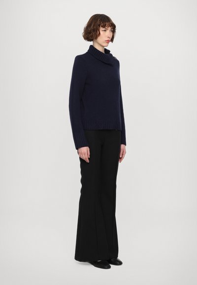 Pull à col roulé marine avec bord côte aux hem et aux poignets, associé à un pantalon évasé noir et des ballerines noires, sur un fond clair uni.
