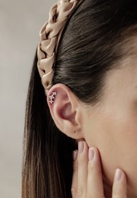 Spleciona satynowa opaska w beżowym kolorze, noszona z srebrną kolczykiem na uchu, ozdobionym wieloma małymi, błyszczącymi kamieniami, na tle ciemnobrązowych włosów.