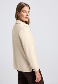 Beżowa bluzka z długim rękawem i kołnierzykiem, gładka w dotyku, z bocznymi rozcięciami i zapięciem na guzik przy mankietach, noszona na ciemno-brązowych spodniach.
