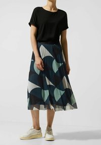 Top corto nero a maniche corte abbinato a una gonna midi plissettata con un motivo geometrico in blu scuro, blu chiaro e crema. Sneakers bianche.