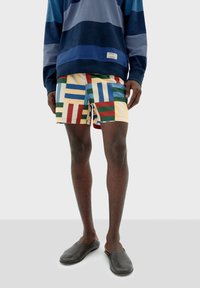 Sudadera de rayas para hombre en azul y gris, combinada con pantalones cortos de baño multicolores con un patrón geométrico. Zapatos oscuros tipo slip-on.