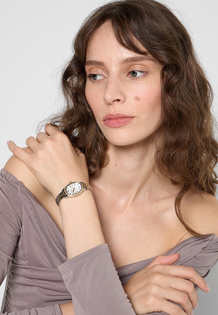 Montre en métal argenté avec un cadran rond blanc, des accents dorés et un bracelet à maillons alternés. Portée au poignet d'une femme avec de longs cheveux.
