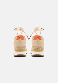 Zapatillas deportivas beige y naranjas con acentos tejidos, que cuentan con una gruesa suela blanca y cordones planos beige, diseñadas para comodidad y soporte.