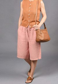 Tan gebreide mouwloze top met knopen, gecombineerd met losse roze linnen shorts en een bruine geperforeerde handtas. Bruin sandalen maken de outfit compleet.