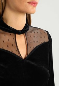 Schwarzes Samtoberteil mit einem transparenten Mesh-Ausschnitt mit gepunktetem Muster und einem Schlüssel-Ausschnitt, ergänzt durch eine schleifenartige Bindung am Kragen.