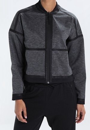 Personne portant une veste zippée gris foncé ajustée avec des bordures noires et un pantalon noir, debout devant un fond uni.