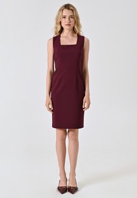 Abito senza maniche color bordeaux con scollo quadrato, taglio su misura, lunghezza al ginocchio e texture liscia. Indossato con tacchi a punta bordeaux.