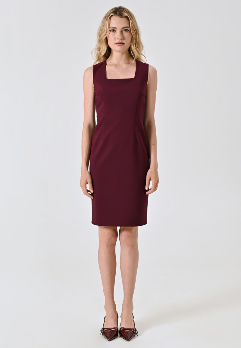 Abito senza maniche color bordeaux con scollo quadrato, taglio su misura, lunghezza al ginocchio e texture liscia. Indossato con tacchi a punta bordeaux.