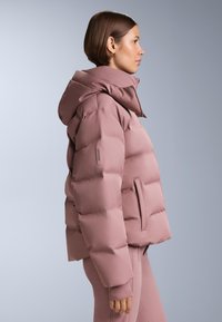 Žena u bočnom profilu nosi jarko ljubičastu prošivenu jaknu s visokim ovratnikom i odgovarajuće ljubičaste hlače na jednostavnoj sivoj pozadini.