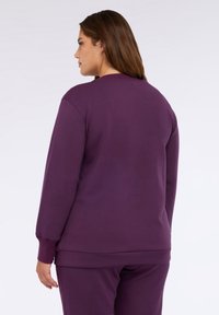 Felpa viola con una vestibilità rilassata, polsini e orlo a coste, e tessuto morbido; indossata con pantaloni coordinati, vista da dietro.