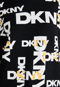 Svart tröja med ett återkommande mönster av texten "DKNY" i vitt och gult, med varierande storlekar och orienteringar på bokstäverna.
