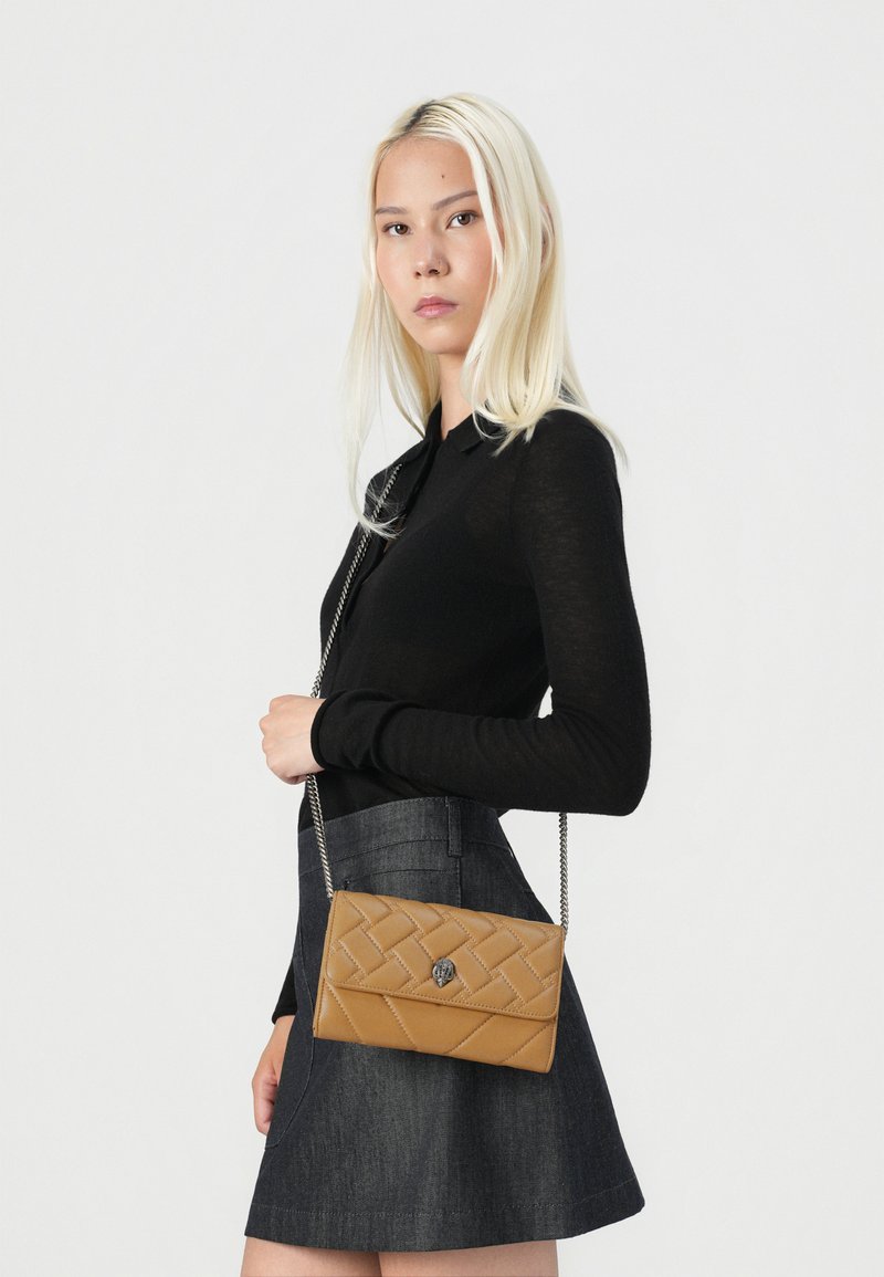 Sac de crossbody matelassé beige avec une texture lisse, une bandoulière en chaîne argentée et un accent logo. Le modèle porte un haut noir à manches longues et une jupe en denim.
