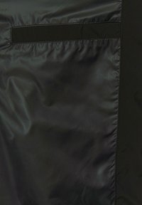 Schwarzer Nylon-Stoff mit glatter Textur, ausgestattet mit einer Tasche oben links und einem dezenten Glanz. Sauberer, minimalistischer Stich entlang der Kanten.