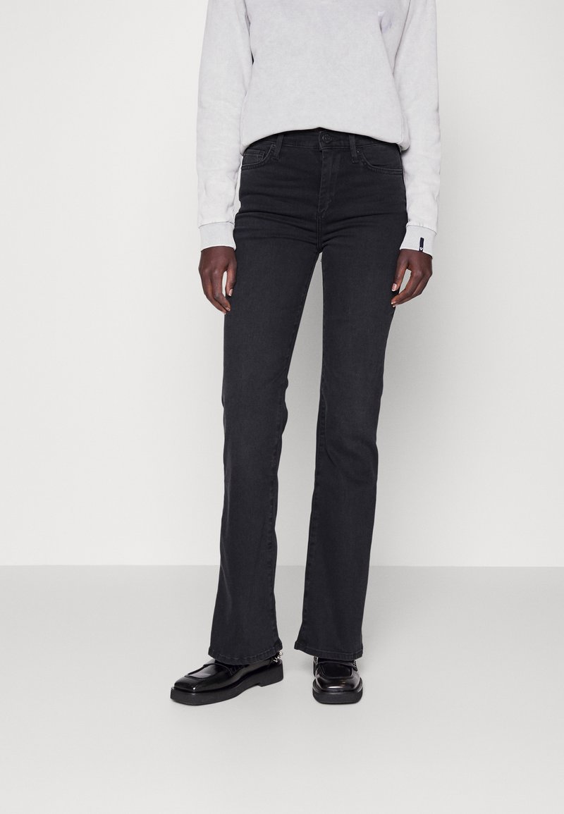 Personne portant un jean noir à jambes droites, des mocassins noirs et un haut à manches longues gris clair, debout devant un fond blanc uni.