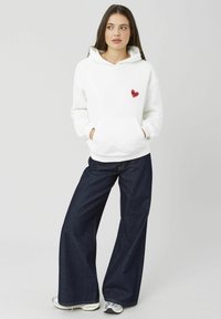 Sweat à capuche blanc avec un dessin de cœur rouge sur le côté gauche de la poitrine, poche avant, poignets côtelés, associé à un jean large foncé.