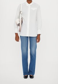 Weißes Hemd mit Kragen und Tasche, kombiniert mit blauen Schlagjeans und schwarzen Ballerinas. Hält eine hellfarbige Umhängetasche.