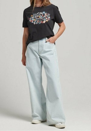 Femme portant un t-shirt noir à imprimé floral rentré dans un jean ample bleu clair, avec des baskets blanches.